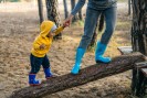 Man sieht ein kleines Kind, das mit Hilfe einer erwachsenen Person, einen schrägen Baumstamm auf einem Spielplatz hochgeht. Das Kind nimmt die Hand des Erwachsenen als Hilfe.
