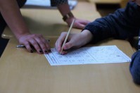 Ausschnittsfotografie von einem Arbeitsblatt auf dem Tisch, man sieht die Hand des Schülers und der Lehrer stützt sich von der anderen Tischseite ab 
